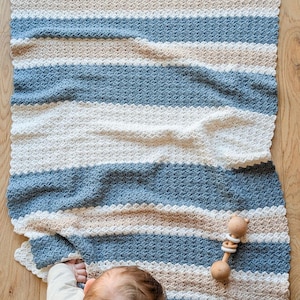 Op de afbeelding: Een handgemaakte, gestreepte babydeken in tinten blauw, beige en wit. De deken is gehaakt met een getextureerde steek en heeft een geschulpte rand. Een houten babyspeelgoed ligt naast de deken.