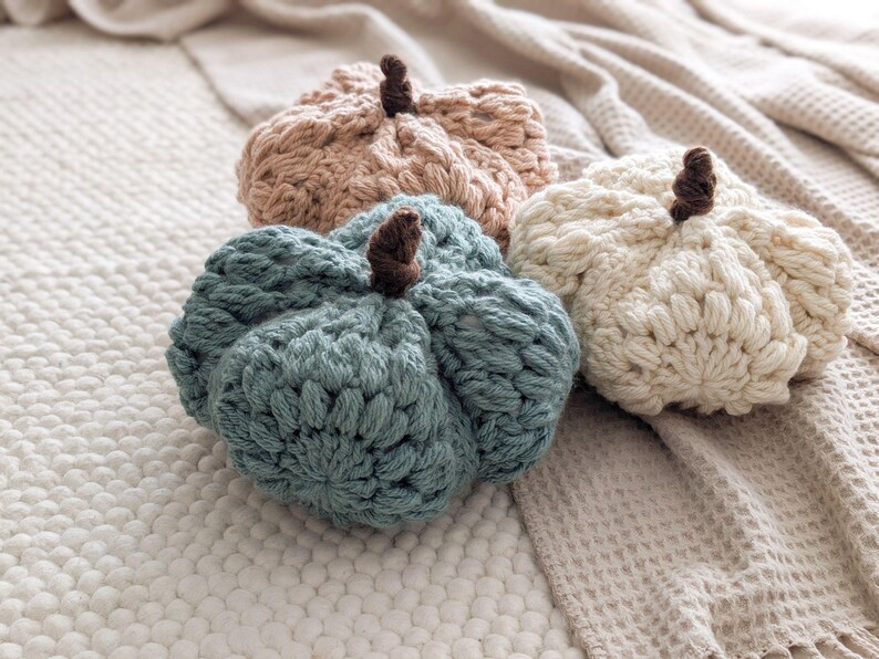 Op de afbeelding: Drie handgemaakte gehaakte pompoenen in gedempte tinten van stoffig blauw, cr&egrave;me en lichtbeige. Elke pompoen heeft een gestructureerd, geribbeld steekpatroon en een kleine, donkerbruine gehaakte steel aan de bovenkant voor een zachte uitstraling.