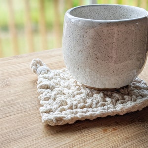 May include: A white ceramic mug with a speckled glaze sits on a crocheted coaster. The coaster is made of natural fiber and has a square shape.