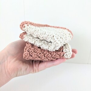 May include: Two crocheted dishcloths, one in a light beige color and the other in a light peach color, are stacked on top of each other. The dishcloths are made with a textured stitch pattern.