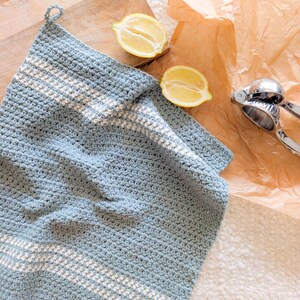 Puede incluir: Una toalla de cocina de crochet azul claro con rayas blancas. La toalla est&aacute; sobre una superficie blanca con dos mitades de lim&oacute;n y un exprimidor de c&iacute;tricos.