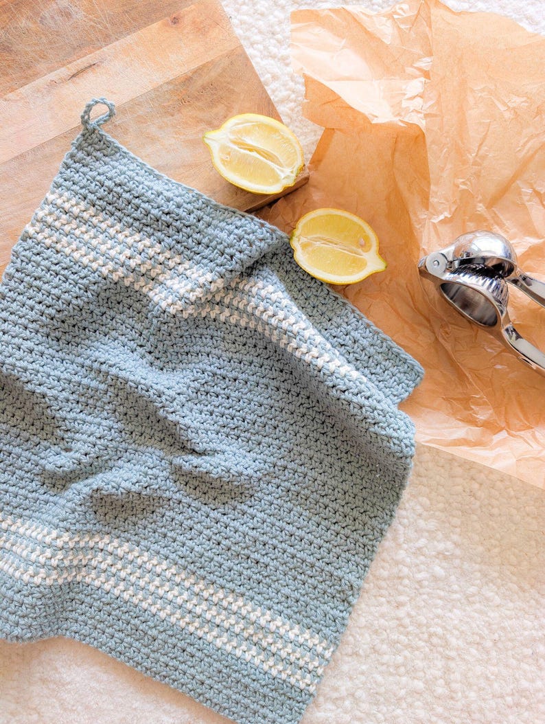 Puede incluir: Una toalla de cocina de crochet azul claro con rayas blancas. La toalla est&aacute; sobre una superficie blanca con dos mitades de lim&oacute;n y un exprimidor de c&iacute;tricos.