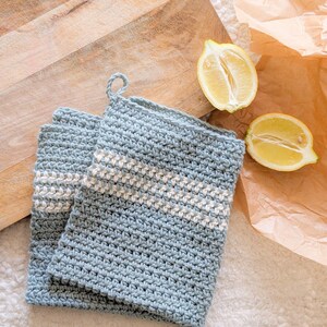 Puede incluir: Dos agarraderas de olla de crochet azul con rayas blancas. Las agarraderas est&aacute;n sobre una superficie blanca con una tabla de cortar de madera y limones en el fondo.