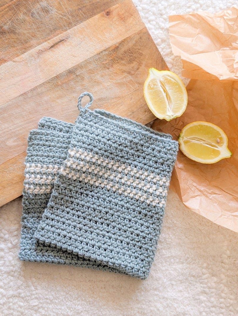 Puede incluir: Dos agarraderas de olla de crochet azul con rayas blancas. Las agarraderas est&aacute;n sobre una superficie blanca con una tabla de cortar de madera y limones en el fondo.