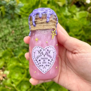 May include: A small, pink potion bottle with a purple, dripped wax seal and a twine-wrapped cork. The bottle has a heart-shaped label with the text "Bella Flores Philter of Love" and gold star accents. The bottle is filled with a shimmering pink liquid.
