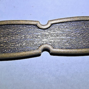 May include: A rectangular, antique-style drawer pull with a dark, textured center and a gold-colored, ornate border. The pull has two holes for mounting and is designed for furniture hardware.