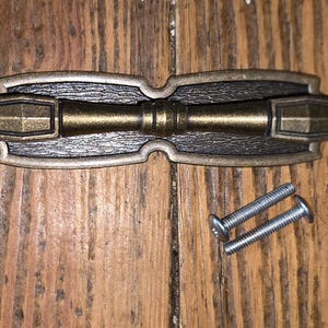 May include: A decorative bronze-toned cabinet pull with a central bar and two end handles. The pull has a textured center section and is accompanied by two silver screws, likely for installation. The hardware is designed for furniture.