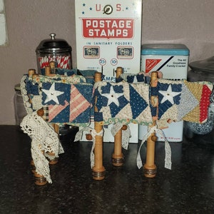May include: Four miniature American flags made of patchwork fabric with a white star on each flag. The flags are attached to wooden dowels with white lace and are displayed on a black countertop.