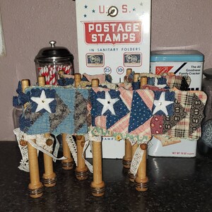 May include: A set of six wooden spools with fabric scraps attached. The fabric scraps are in shades of blue, red, white, and black. The spools are decorated with lace and have a vintage look.