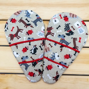 May include: Two heart-shaped oven mitts with a tan background covered in dog illustrations, bones, and fire hydrants. The mitts are trimmed with red piping.