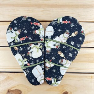 Puede incluir: Dos manoplas de horno con un fondo negro que presenta un estampado caprichoso de muñecos de nieve. Los muñecos de nieve llevan sombreros y bufandas, y están rodeados de copos de nieve y pájaros. Las manoplas están ribeteadas con ribetes verdes.