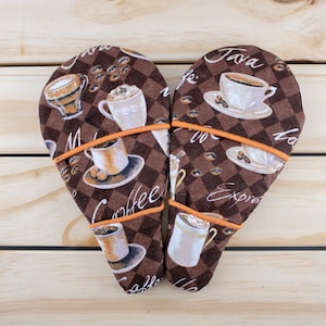 Puede incluir: Dos manoplas de horno, dispuestas en forma de corazón, con un estampado a cuadros marrón y blanco con ilustraciones de tazas de café y granos de café. Las manoplas tienen ribetes naranjas y las palabras "Coffee", "Java" y "Espresso".