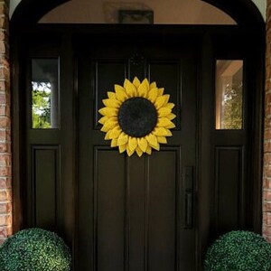 Puede incluir: Una puerta delantera negra con una corona de girasol amarilla. La puerta tiene un panel de vidrio a cada lado y una manija de puerta negra. Hay dos arbustos verdes en macetas negras a cada lado de la puerta.