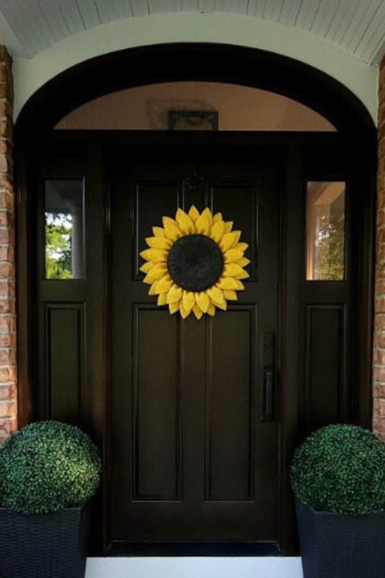 Puede incluir: Una puerta delantera negra con una corona de girasol amarilla. La puerta tiene un panel de vidrio a cada lado y una manija de puerta negra. Hay dos arbustos verdes en macetas negras a cada lado de la puerta.