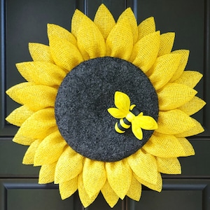 Puede incluir: Una corona de girasol amarilla con un centro negro y una peque&ntilde;a decoraci&oacute;n de abeja. La corona est&aacute; hecha de arpillera y tiene un aspecto r&uacute;stico.