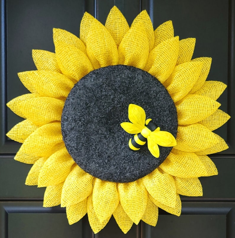 Puede incluir: Una corona de girasol amarilla con un centro negro y una peque&ntilde;a decoraci&oacute;n de abeja. La corona est&aacute; hecha de arpillera y tiene un aspecto r&uacute;stico.