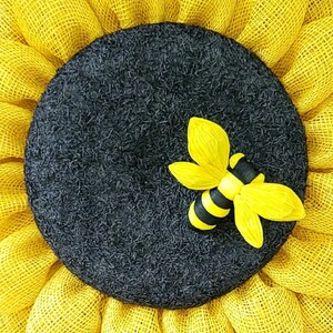 Puede incluir: Una corona de girasol de arpillera amarilla con un centro negro y una aplicaci&oacute;n de abeja amarilla y negra.