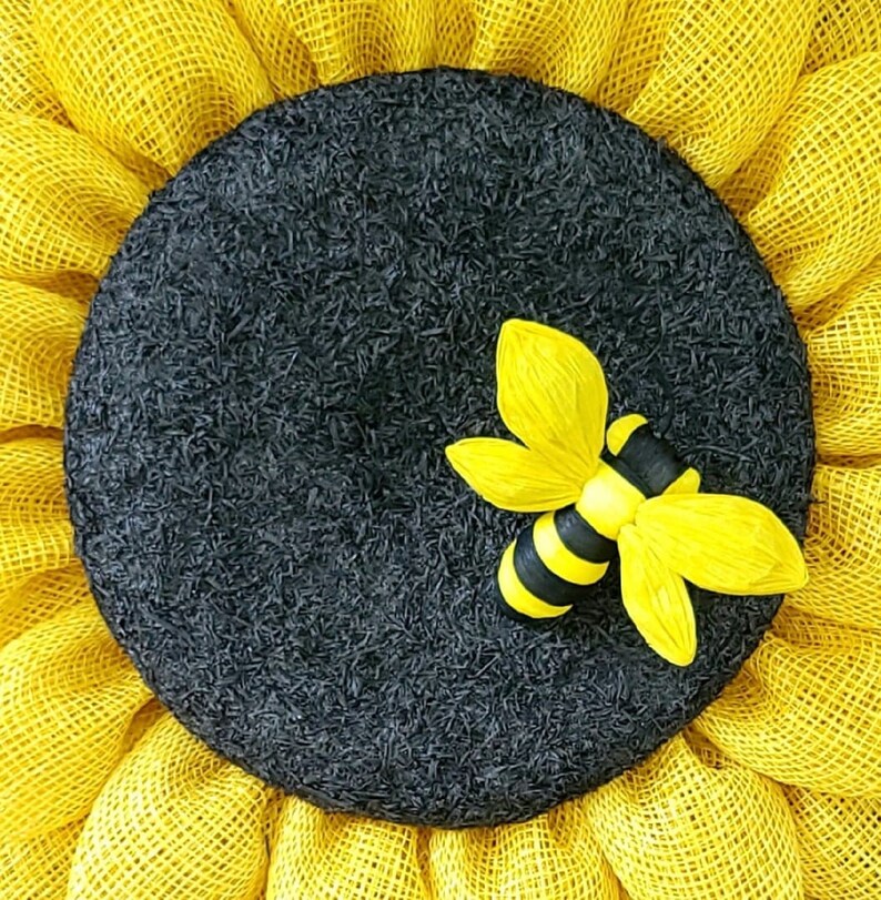 Puede incluir: Una corona de girasol de arpillera amarilla con un centro negro y una aplicaci&oacute;n de abeja amarilla y negra.