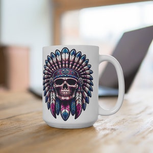 May include: White ceramic mug featuring a detailed illustration of a skull wearing a Native American headdress. The headdress is adorned with blue, pink, and purple feathers. The mug has a large handle and is set on a wooden surface.