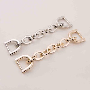 Puede incluir: Dos correas de cadena metálicas, una plateada y otra dorada, cada una con una hebilla en forma de D en ambos extremos. Las cadenas están compuestas por eslabones ovalados. La cadena dorada tiene un acabado cálido y brillante, mientras que la plateada tiene un tono más frío.