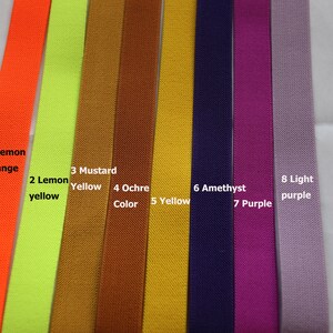 May include: Eight different colours of elastic ribbon, from left to right: lemon orange, lemon yellow, mustard yellow, ochre, yellow, amethyst, purple, and light purple. The ribbon is labelled with the colour name and number.