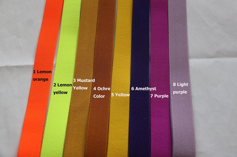 May include: Eight different colours of elastic ribbon, from left to right: lemon orange, lemon yellow, mustard yellow, ochre, yellow, amethyst, purple, and light purple. The ribbon is labelled with the colour name and number.