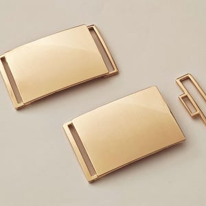 Könnte beinhalten: Zwei goldfarbene Metall-Gürtelschließen. Eine ist eine rechteckige Schließe mit glatter Oberfläche, die andere ist eine kleinere, rechteckige Schlaufe.