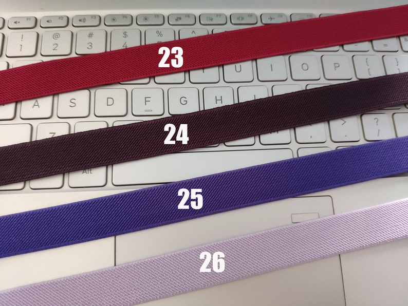 May include: Four different colors of elastic bands, red, brown, purple, and light purple, are laid out on a white keyboard. The bands are numbered 23, 24, 25, and 26.