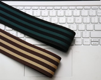 May include: Two striped ribbons, one green and black, the other brown and tan, rest on a laptop keyboard. The ribbons have a ribbed texture and are likely for crafting or sewing projects. The keyboard is white.