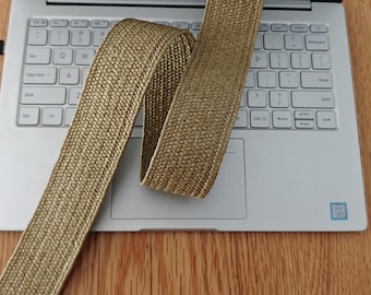 May include: A long, continuous strip of woven elastic webbing in a metallic gold tone. The material features a dense, textured, braided pattern throughout its length, showcasing a consistent weave and a slight sheen across the surface of the fabric.