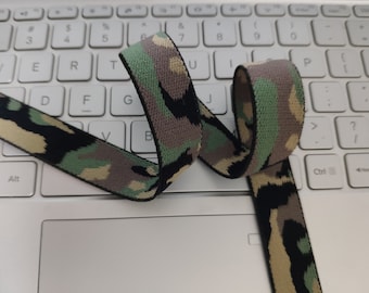 May include: Camouflage patterned ribbon in shades of green, tan, black, and gray, coiled on a white laptop keyboard. The ribbon's design resembles military camouflage, with a woven texture.