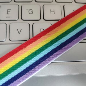May include: Rainbow striped elastic band on a white keyboard. The band is made of woven fabric and has red, orange, yellow, green, blue, indigo, and violet stripes.