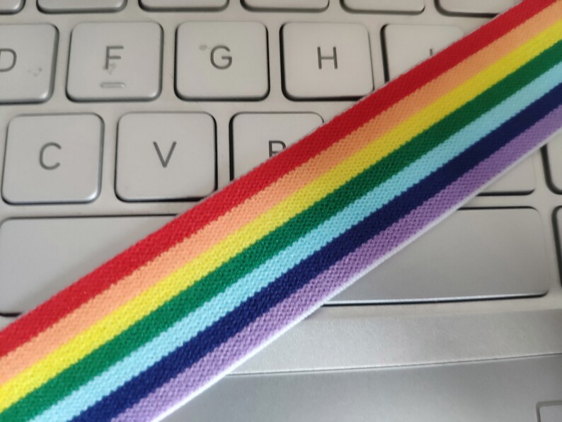 May include: Rainbow striped elastic band on a white keyboard. The band is made of woven fabric and has red, orange, yellow, green, blue, indigo, and violet stripes.