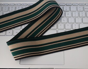 May include: A length of striped ribbon in green, tan, and black, resting on a laptop keyboard. The ribbon has a repeating pattern of stripes, with the green stripes being the most prominent. The ribbon is likely used for crafting or sewing projects.