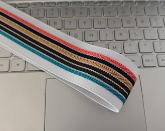 May include: White elastic band with a colorful chevron pattern in shades of teal, brown, gold, black, and coral. The band is lying on a laptop keyboard.