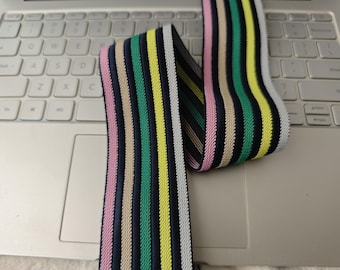 May include: A colorful striped elastic band featuring vertical stripes in pink, green, yellow, beige, and white against a navy blue background. The elastic is shown on a laptop keyboard, highlighting its flexibility and potential use in various crafts and sewing projects.