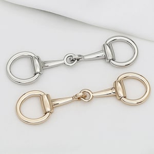 Pode incluir: Dois freios de cavalo de metal, um prateado e outro dourado, são exibidos sobre um fundo branco. Cada freio apresenta um anel em D em cada extremidade e uma articulação central. O freio dourado tem um acabamento quente e polido, enquanto o prateado tem uma superfície brilhante e reflexiva.