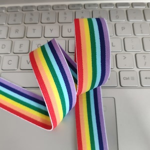 May include: Rainbow striped elastic band with white edges. The band is folded in half and lying on a white laptop keyboard.