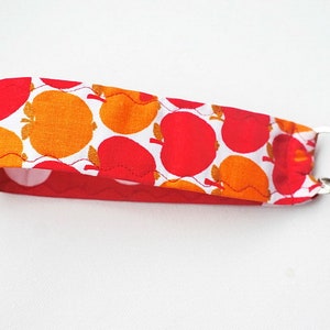 Könnte beinhalten: Ein Schlüsselanhänger aus rotem und orangefarbenem Stoff mit weißem Hintergrund und einem Muster aus Äpfeln. Der Schlüsselanhänger hat einen silbernen Schlüsselring und eine rote Perle.