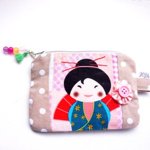 Könnte beinhalten: Eine beige Reißverschlusstasche mit weißem Punktmuster. Die Tasche zeigt eine bunte Applikation einer japanischen Puppe mit einem rot-blauen Kimono und einer rosa Blume mit einem Knopf.
