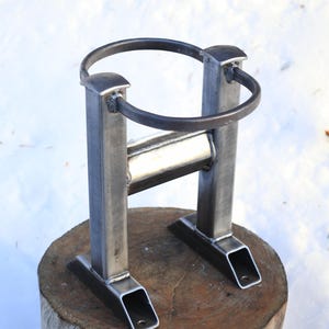 Peut inclure: Support de foyer en m&eacute;tal fait &agrave; la main. Le support est en acier soud&eacute; avec un anneau sup&eacute;rieur circulaire et une base rectangulaire. Il est con&ccedil;u pour maintenir un anneau de foyer et est pr&eacute;sent&eacute; sur une base en bois.