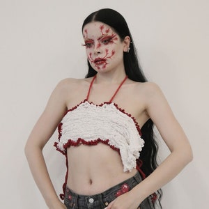 May include: A woman with long black hair and red makeup on her face is wearing a white crocheted halter top with red trim and dark denim jeans with red paint splatters.