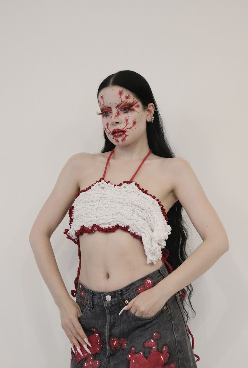 May include: A woman with long black hair and red makeup on her face is wearing a white crocheted halter top with red trim and dark denim jeans with red paint splatters.