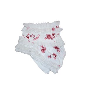 Könnte beinhalten: Weiße Baby-Bloomers mit Rüschenbesatz und einem Blumenmuster in Rottönen. Die Bloomers haben ein mehrschichtiges Design mit mehreren Reihen von Rüschen. Das Blumenmuster ist über den Stoff verstreut.