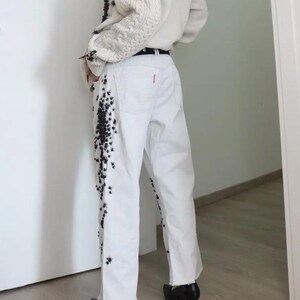 May include: White denim jeans featuring a black floral or insect-like embellishment running down the outer side of the legs. The jeans have a straight-leg cut, a raw hem at the ankles, and a small red tab detail on the back pocket area.
