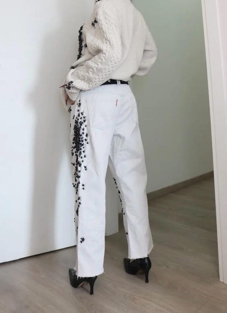 May include: White denim jeans featuring a black floral or insect-like embellishment running down the outer side of the legs. The jeans have a straight-leg cut, a raw hem at the ankles, and a small red tab detail on the back pocket area.