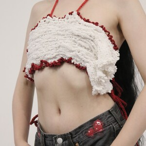 May include: A white crocheted halter top with a red crocheted trim. The top is worn over a pair of dark denim jeans.