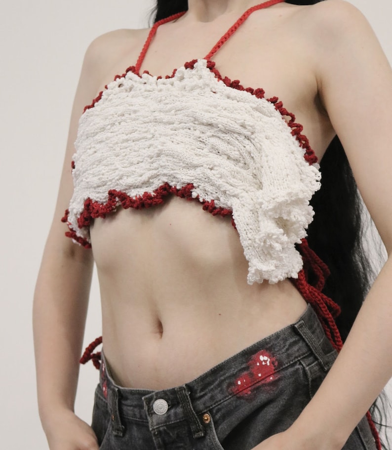 May include: A white crocheted halter top with a red crocheted trim. The top is worn over a pair of dark denim jeans.
