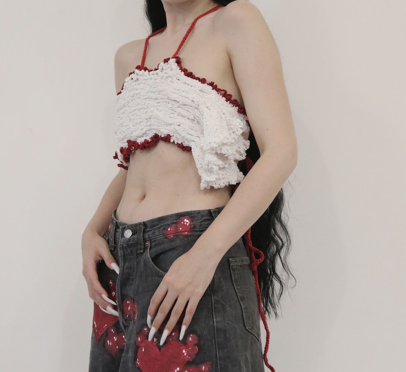May include: A white crochet halter top with red trim and a ruffled hem. The top is worn over a pair of dark gray jeans with red paint splatters.