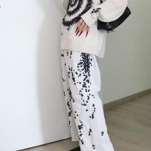 May include: An off-white cable knit sweater featuring a large black spiral pattern and matching black embellishments, paired with white wide-leg trousers decorated with scattered black floral-like embroidery, a black shoulder bag, and black heels.